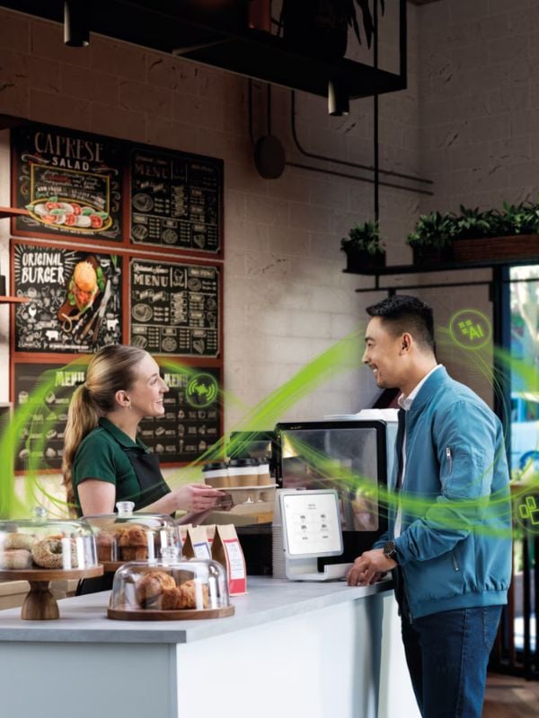 Man smiling at a barista in a café, engaging in conversation, with a display of pastries and a digital ordering kiosk in view, illustrating a friendly customer service experience enhanced by technology.